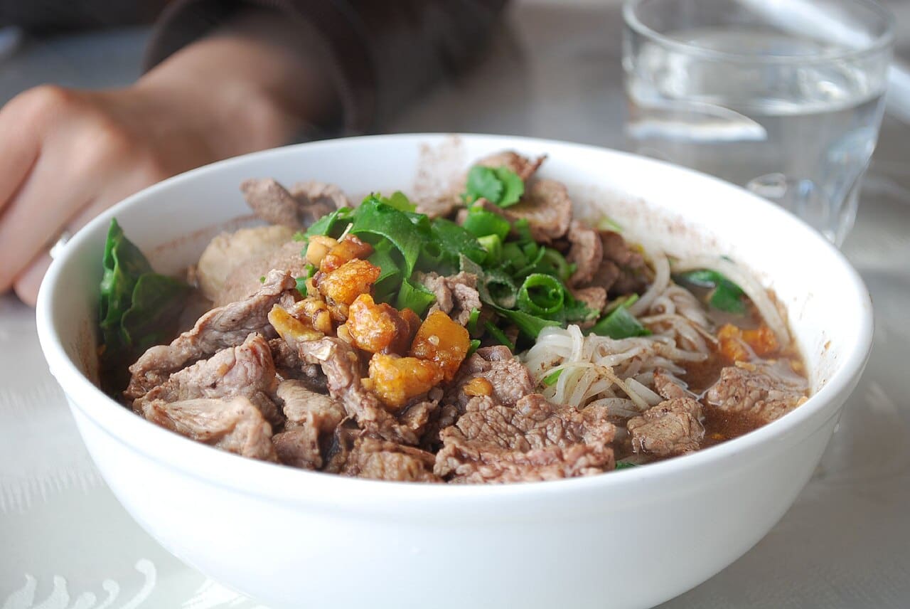 A bowl of Thai boat noodles with dark broth, pork, herbs and bean sprouts